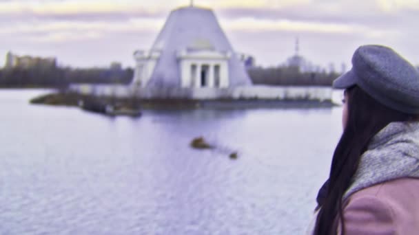 Jeune femme pensive en manteau rose debout au bord du lac regardant de côté, effet caméra VHS. Images d'archives. Jeune fille aux cheveux longs foncés debout près de l'eau en saison froide . — Video