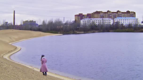 Mujer morena joven con abrigo cálido rosa caminando sola cerca del lago en otoño, efecto VHS. Imágenes de archivo. Chica solitaria caminando sobre la arena en tiempo frío . — Vídeos de Stock