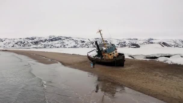 Opuštěná stará loď na písčitém pobřeží u moře se zasněženými kopci. Záběry. Letecký pohled na krásnou zimní krajinu s troskami lodi, lidé kráčející po břehu a moře. — Stock video