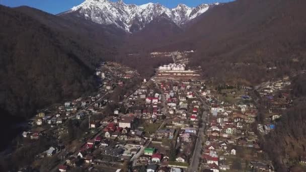 Veduta aerea di una piccola città con catena montuosa con cima innevata e cielo azzurro chiaro sullo sfondo. Clip. Volare sul villaggio situato tra le montagne con pendii boscosi . — Video Stock