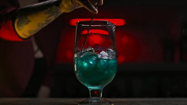 Close-up of bartender stirring cocktail with ice with spoon. Stock footage. Beautiful mixing with cocktail spoon of colored drink with ice on dark neon background — Stock Photo, Image