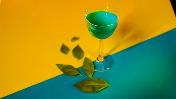 Ustawienie napoju alkoholowego na wielokolorowym tle. Materiał filmowy. Niebieski napój alkoholowy stoi na dekoracyjnym kolorowym tle z zielonymi liśćmi zdobiącymi tło — Wideo stockowe