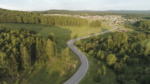 Letecká prázdná cesta lesem, letní přírodní krajina. Scéna. Létání nad dálnicí přes zelený smrkový les s modrou zataženou oblohou a malou vesničkou na pozadí. — Stock fotografie