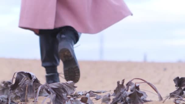 Rear view of female black shoes on the sand, close up view. Stock footage. Woman wearing black leather boots and pink coat walking away on sandy coast with dry tree leaves. — Stock Video