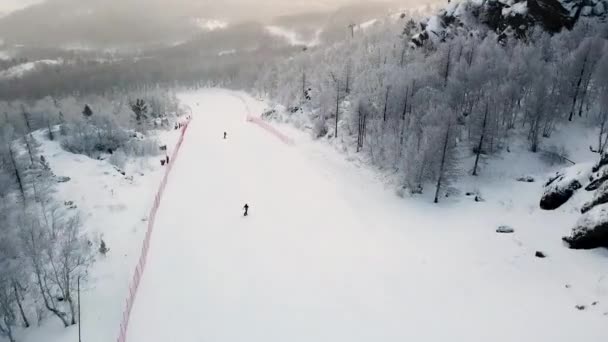 Flygfoto över skidorten och snöiga berg täckt av barrskog. Lagerbilder. Vit backe med skidspår och snowboard människor, sport och natur koncept. — Stockvideo