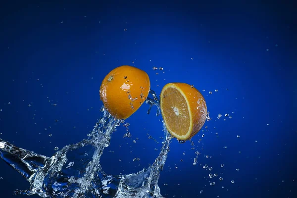Dos mitades de naranja y salpicadura de agua sobre fondo azul —  Fotos de Stock
