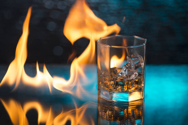 glass of whisky with ice on a blue background and fire flames