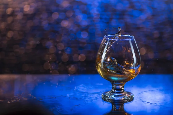 Bicchiere da vino di cognac e spruzzi su uno sfondo blu bokeh — Foto Stock