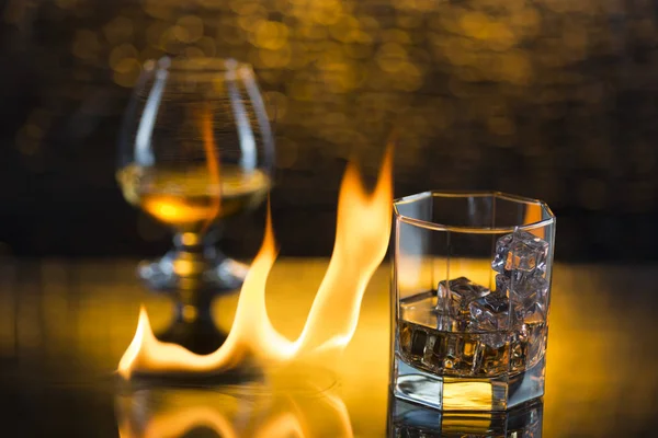 Glass o whisky with ice and wineglass of cognac and fire flames on yellow bokeh background — Stock Photo, Image