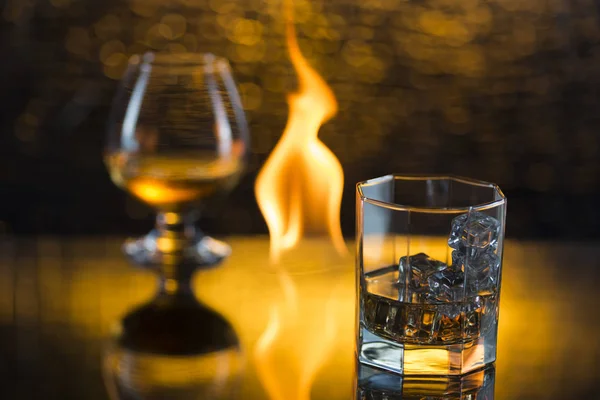 Glass o whisky with ice and wineglass of cognac and fire flames on yellow bokeh background — Stock Photo, Image