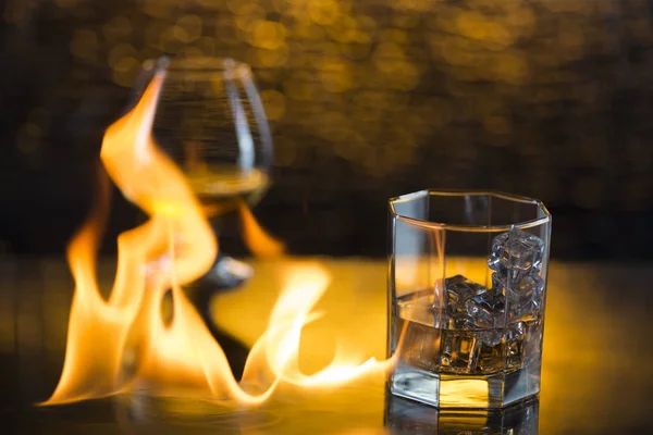 Vaso o whisky con hielo y vino de coñac y llamas de fuego sobre fondo amarillo bokeh — Foto de Stock