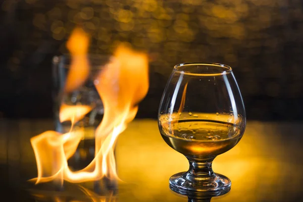Bicchiere o whisky con ghiaccio e bicchiere da vino di cognac e fiamme di fuoco su sfondo giallo bokeh — Foto Stock