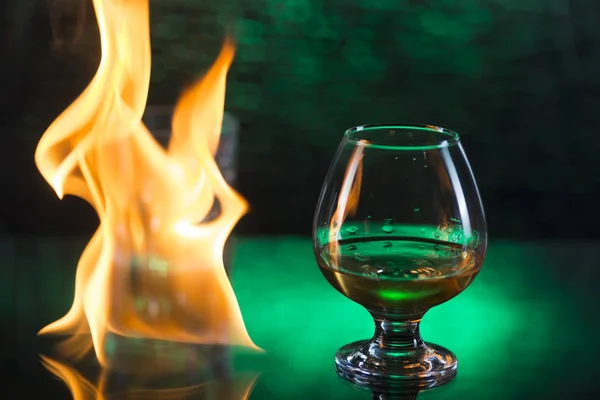 Vaso o whisky con hielo y vino de coñac y llamas de fuego sobre fondo verde bokeh — Foto de Stock