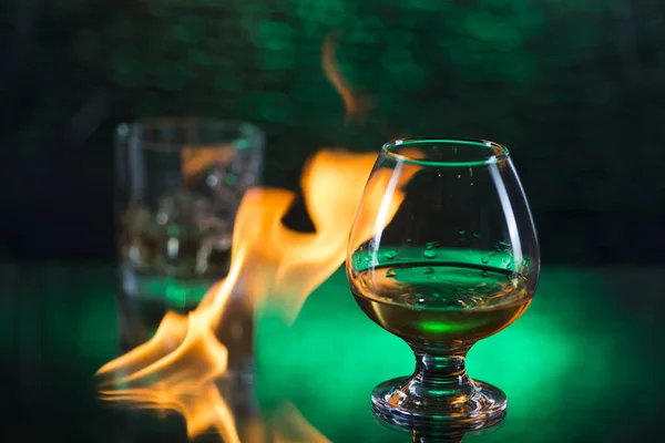 O viski bardağı konyak ve buz ile cam ve yeşil bokeh arka plan üzerinde alevler ateş — Stok fotoğraf