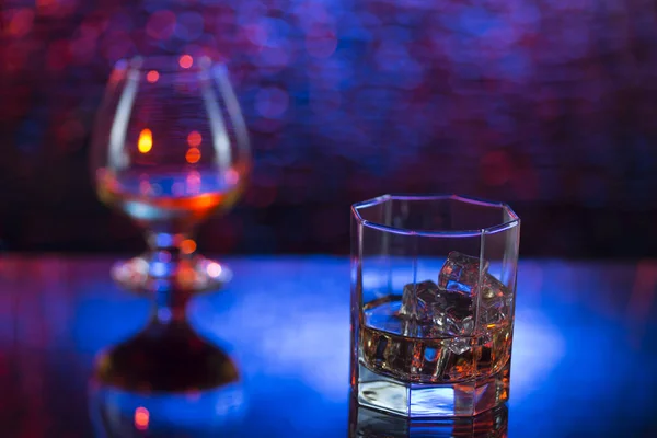 Bicchiere o whisky con ghiaccio e bicchiere da vino di cognac su sfondo bokeh rosa-blu — Foto Stock