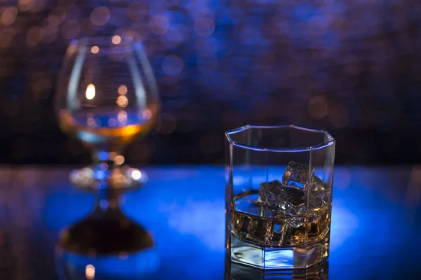 Bicchiere o whisky con ghiaccio e bicchiere da vino di cognac su sfondo bokeh giallo-blu — Foto Stock