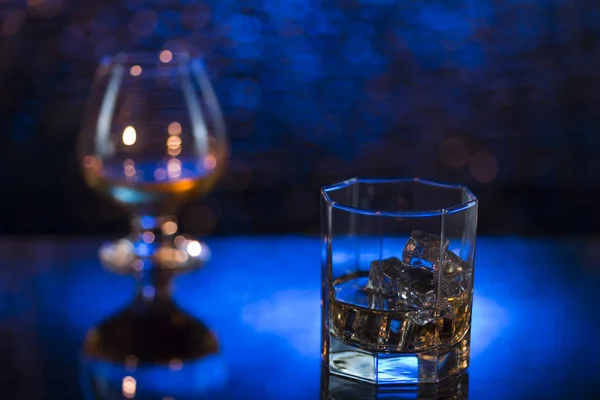 Bicchiere o whisky con ghiaccio e bicchiere da vino di cognac su sfondo blu bokeh — Foto Stock