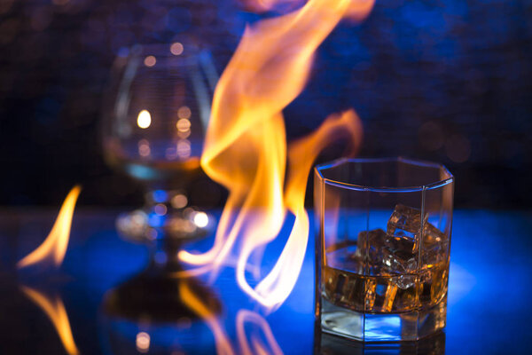 Glass o whisky with ice and wineglass of cognac and fire flames on bokeh background