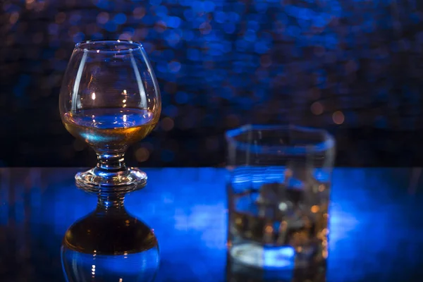 Bicchiere o whisky con ghiaccio e bicchiere da vino di cognac su sfondo blu bokeh — Foto Stock