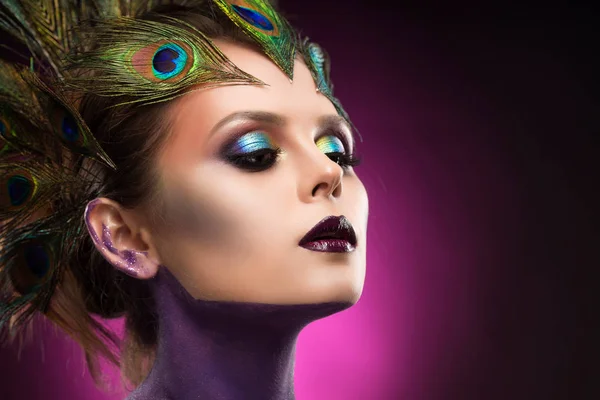 Menina de peito grande bonita vestindo penas de pavão em seu cabelo e artístico violeta corpo brilhante arte em seu pescoço, maquiagem vanguarda — Fotografia de Stock