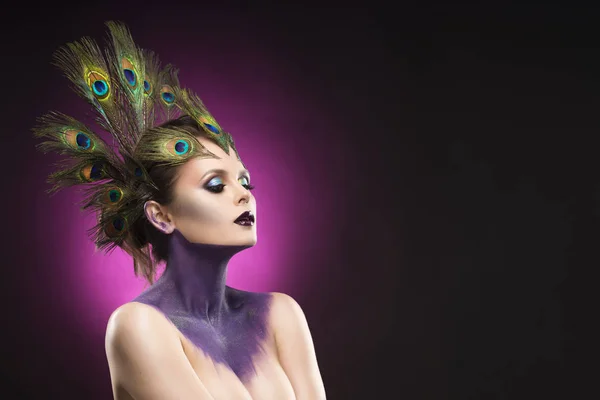 Schöne große Brust Mädchen trägt Pfauenfedern im Haar und künstlerische violett glänzende Körperkunst am Hals, Avantgarde-Make-up — Stockfoto