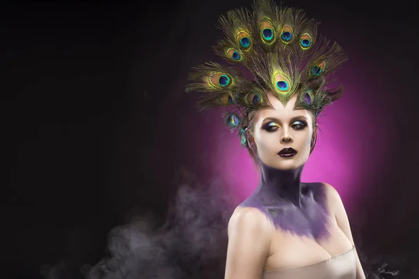 Beautiful big breast girl wearing peacock feathers in her hair and artistic violet shiny body art on her neck, vanguard makeup in theatrical smoke Stock Photo
