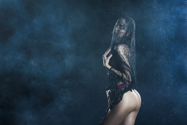 Gothic style young beautiful girl wearing a red lingerie and black veil on her head and face sensually posing in water drops and shows her butt in a theatrical smoke. Healthy smooth skin. Copy space