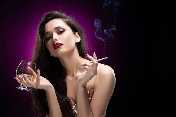 肉感的輝くドレスを着ている美しい魅力的なブルネットの少女は 彼女の手で氷とタバコ喫煙の煙でウィスキーのグラスを保持しています 赤い唇にきらめく 黒紫色の背景 — ストック写真