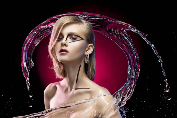 Beautiful blonde girl with naked shoulders and avant-garde conceptual makeup, surrounded by splashes and drops of water on a black and red background. Copy space. Advertising and commercial design.