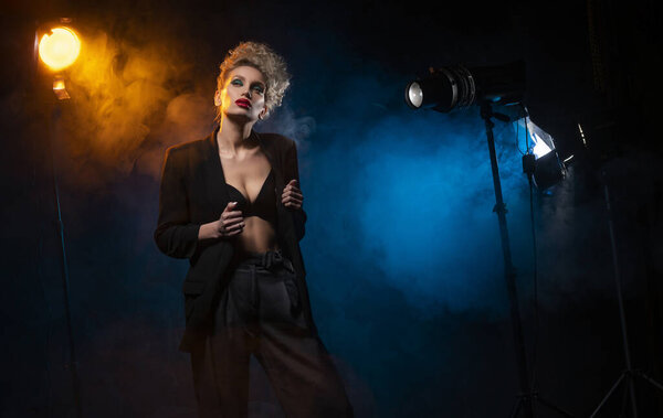 A beautiful blonde girl with an elegant hairstyle and large breasts, wearing a bra, trousers and a blazer, artistically poses in the rays of spotlights in the smoke. Cinematic, art, commercial design