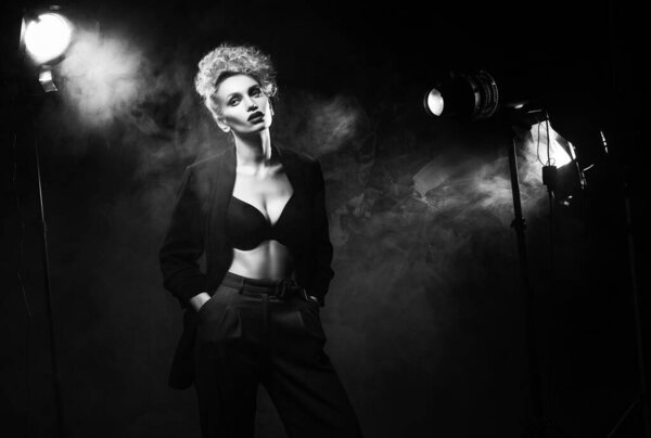 A beautiful blonde girl with an elegant hairstyle and large breasts, wearing a bra, trousers and a blazer, artistically poses in the rays of spotlights in the smoke. Cinematic, art, monochrome design