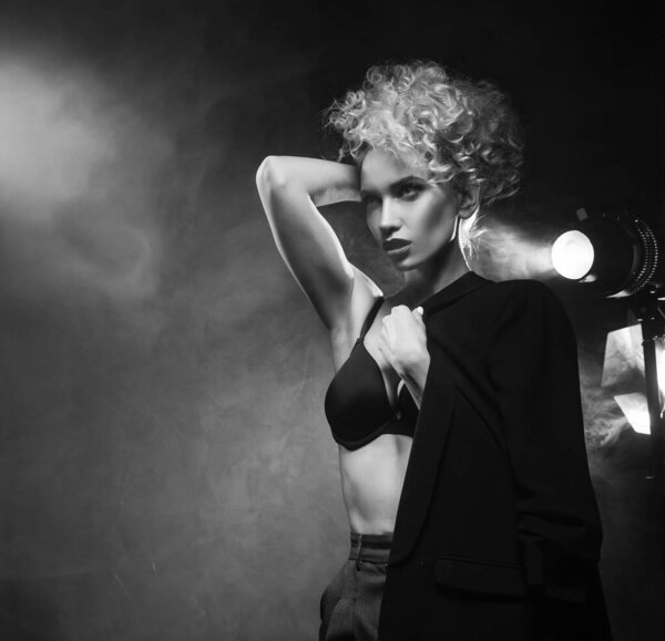 A beautiful blonde girl with an elegant hairstyle and large breasts, wearing a bra, trousers and a blazer, artistically poses in the rays of spotlights in the smoke. Cinematic, art, monochrome design