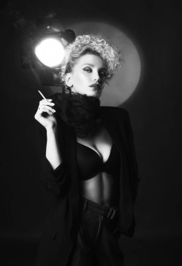 Zarif bir saç stili ve büyük göğüsleri olan güzel bir sarışın kız, sütyen, pantolon, atkı ve ceket giyiyor, spot ışıklarının yanında sigara içiyor. Moda, monokrom, kara film tasarımı