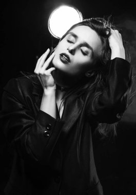 Kırmızı dudaklı, siyah ceketli güzel, ince bir kız model, koyu arka planda eli ile saçına dokunuyor, bir lambanın yanında poz veriyor. Reklamcılık, moda, tek renkli tasarım