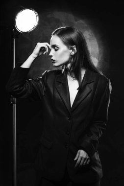 Kırmızı dudaklı, siyah ceketli güzel, ince bir kız model, elini koyu arkaplana koyup yüzüne dokunuyor, bir lambanın yanında poz veriyor. Reklamcılık, monokrom, siyah beyaz tasarım
