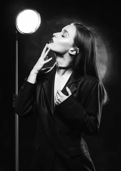 Beautiful slim girl model with red lips, wearing a black blazer, touches her face with her hand on a dark background, posing next to a light lamp. Advertising, monochrome, black and white design