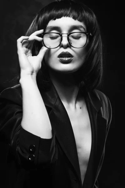 美丽的无褐色纤细女孩 身穿未扣子的黑色夹克 眼镜和深色背景的黑发假发 坐在灯光下 艺术设计 — 图库照片