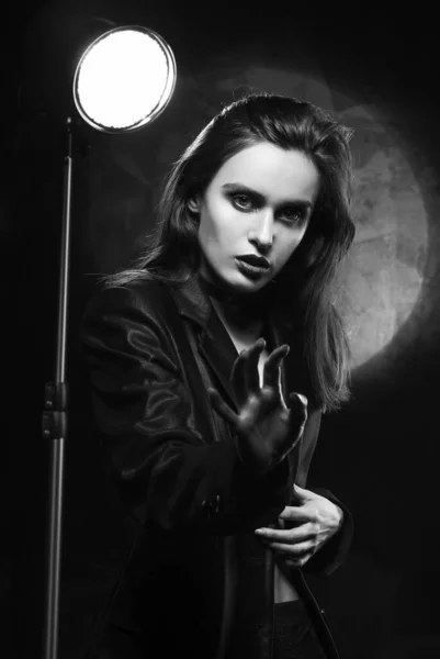 Beautiful slim girl model with red lips, wearing a black blazer, fearfully protects herself with her hand on a dark background, next to the light lamp. Advertising, trendy, monochrome design