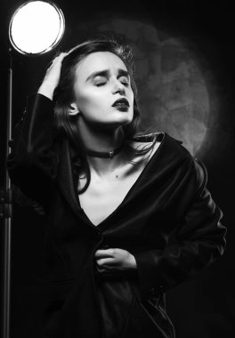 Güzel, ince sutyensiz bir kız, düğmesiz siyah bir ceket giyiyor, şehvetli bir şekilde kaşlarını çatıyor ve koyu bir arka planda eli ile saçlarına dokunuyor, açık bir lambanın yanında poz veriyor. Reklam monokrom tasarımı