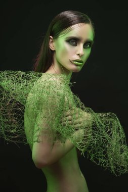 A beautiful slim topless girl with avant-garde makeup covers her breast with green net on a black background. Artistic, fashionable, advertising design clipart