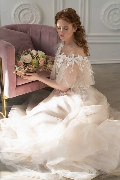 Beautiful natural redhead girl bride, with nude makeup, wearing a white dress, holds a wedding bouquet in her hands and sits on the floor at the chair in a light interior