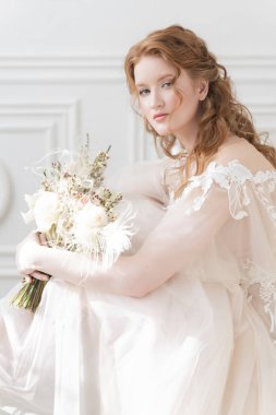 Beyaz bir elbise giymiş, çıplak makyajlı, güzel, kızıl saçlı bir gelin elinde bir düğün buketi tutuyor ve açık bir iç mekânda sandalyede oturuyor.