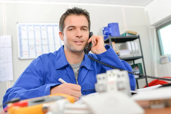 Uomo in tuta da lavoro al telefono, seduto alla scrivania — Foto Stock