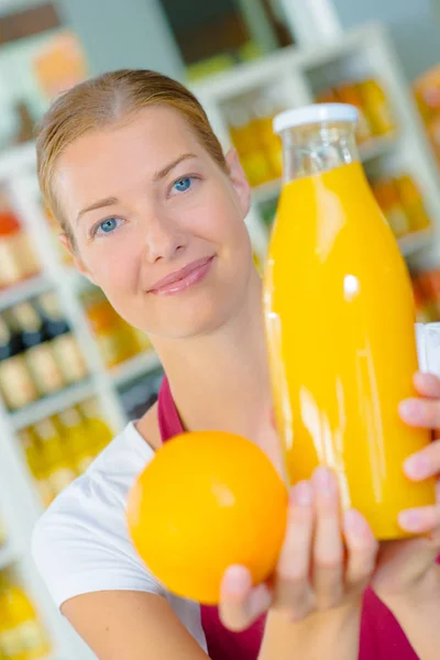 Asistente de tienda sosteniendo una naranja y una botella de jugo de naranja — Foto de Stock