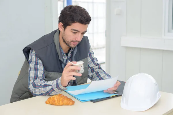 Hombre desayunando y mirando el papeleo en la oficina del sitio —  Fotos de Stock
