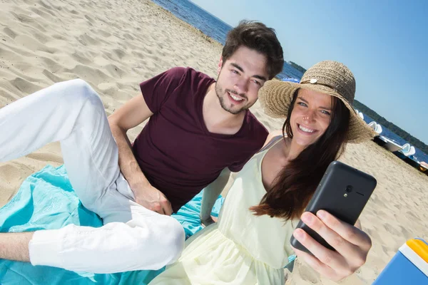 Lindo jovem casal sentado na praia e fazendo selfie — Fotografia de Stock