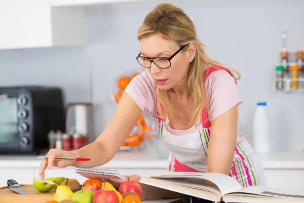 Mogen kvinna läser kokbok i köket letar recept — Stockfoto