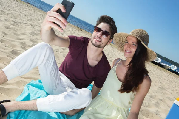 Lindo jovem casal sentado na praia e fazendo selfie — Fotografia de Stock