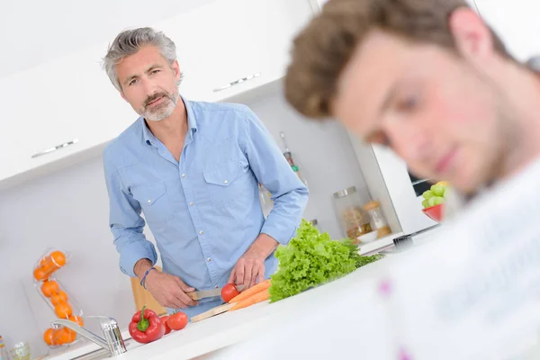 Man matlagning, tittar mot yngre man håller bok — Stockfoto