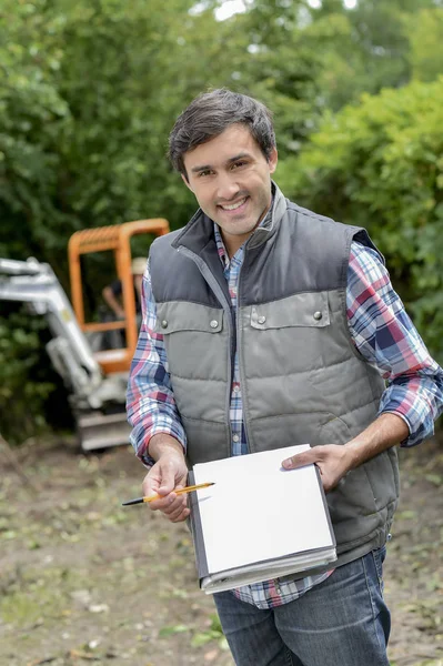 Man outside in front of digger holding paper and folder — Stock Photo, Image